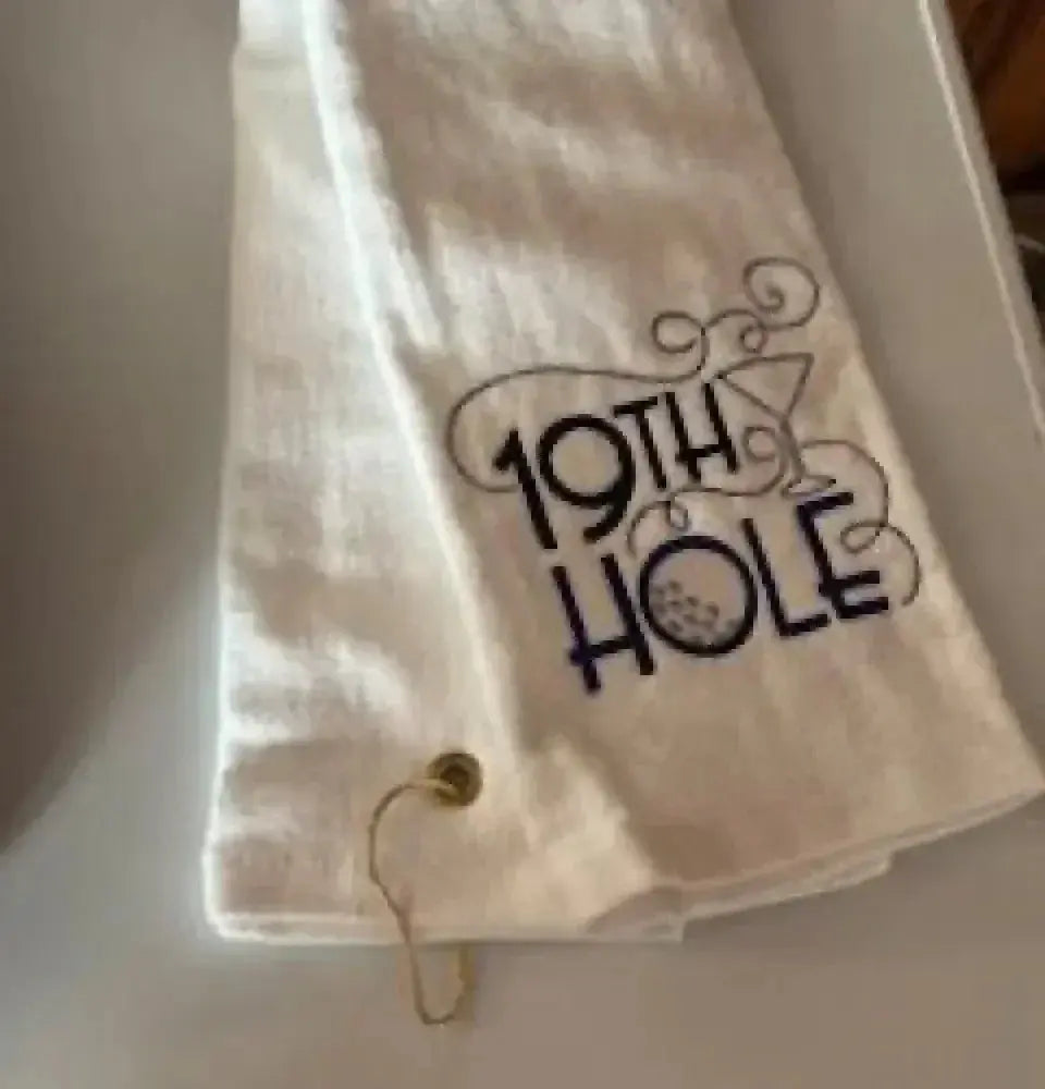 White tri-fold golf towel with grommet, embroidered with '19th Hole' and martini glass design