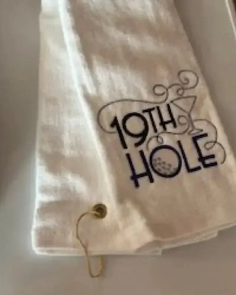 White tri-fold golf towel with grommet, embroidered with '19th Hole' and martini glass design