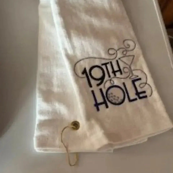 White tri-fold golf towel with grommet, embroidered with '19th Hole' and martini glass design