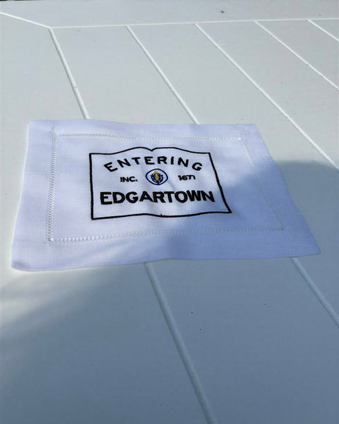 White fabric cocktail napkin with Entering Edgartown INC. 1671 embroidered in black on a white slatted table.
