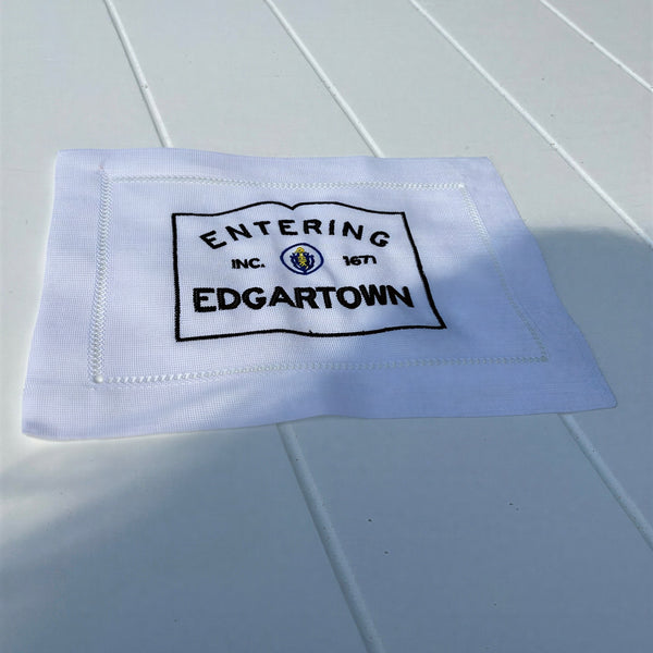 White fabric cocktail napkin with Entering Edgartown INC. 1671 embroidered in black on a white slatted table.
