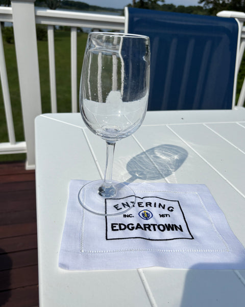 Clear glass on a white fabric cocktail napkin with black Entering Edgartown embroidery on a white outdoor table.