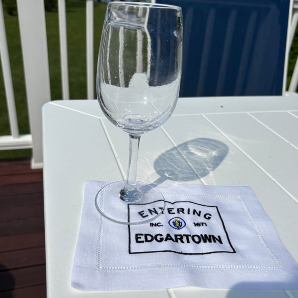 Clear glass on a white fabric cocktail napkin with black Entering Edgartown embroidery on a white outdoor table.