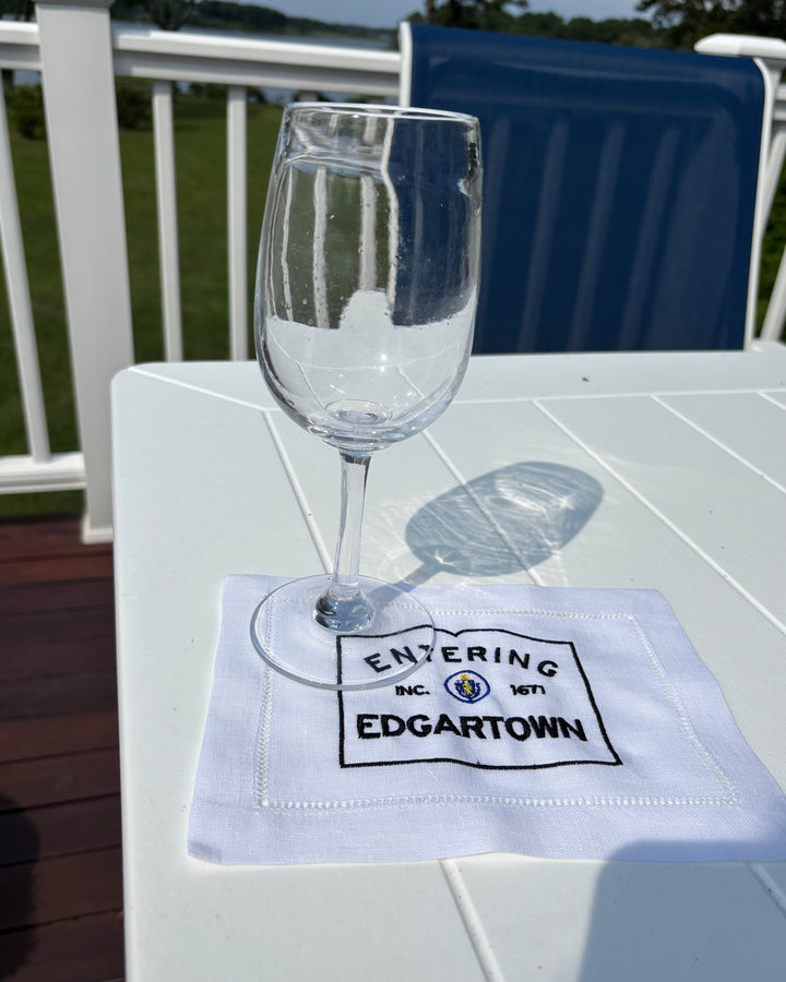 Clear glass on a white fabric cocktail napkin with black Entering Edgartown embroidery on a white outdoor table.