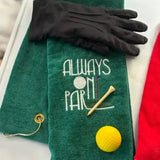 Green embroidered golf towel with 'Always On Par' text, golf tee, yellow golf ball, and black glove