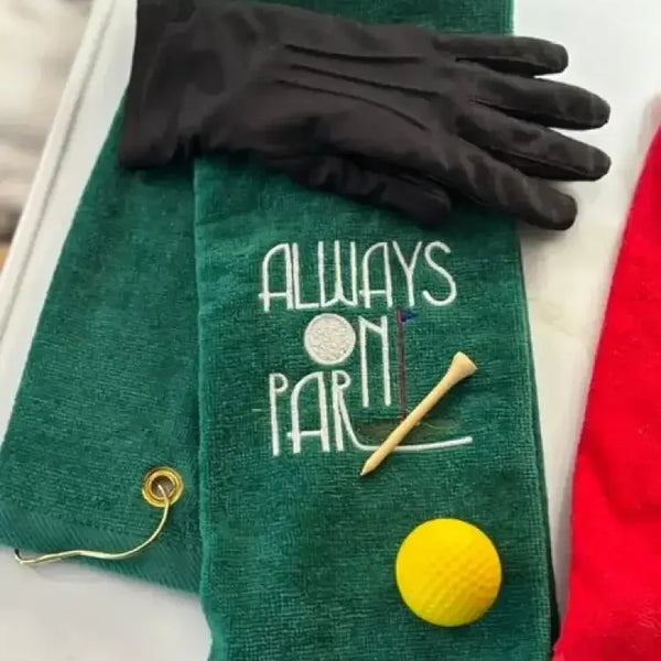 Green embroidered golf towel with 'Always On Par' text, golf tee, yellow golf ball, and black glove