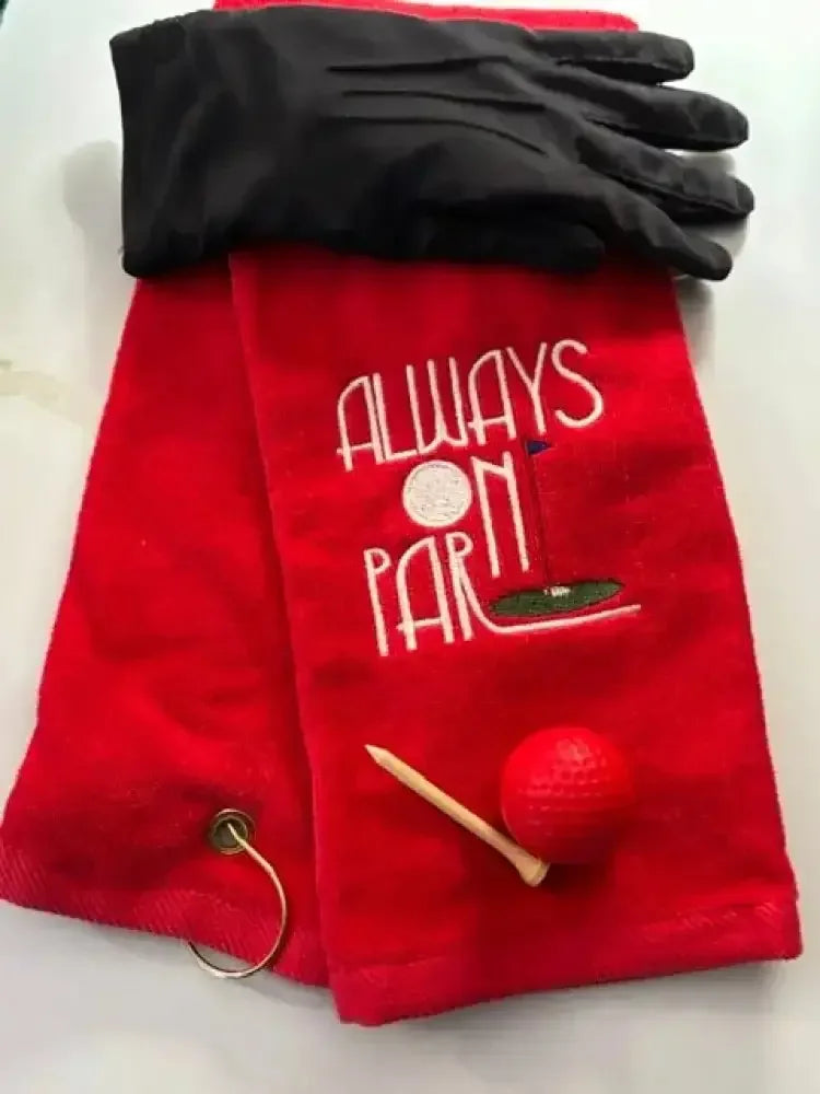 Red embroidered golf towel with 'Always On Par' text, black golf glove, tee, and red golf ball