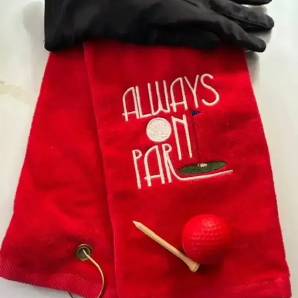 Red embroidered golf towel with 'Always On Par' text, black golf glove, tee, and red golf ball