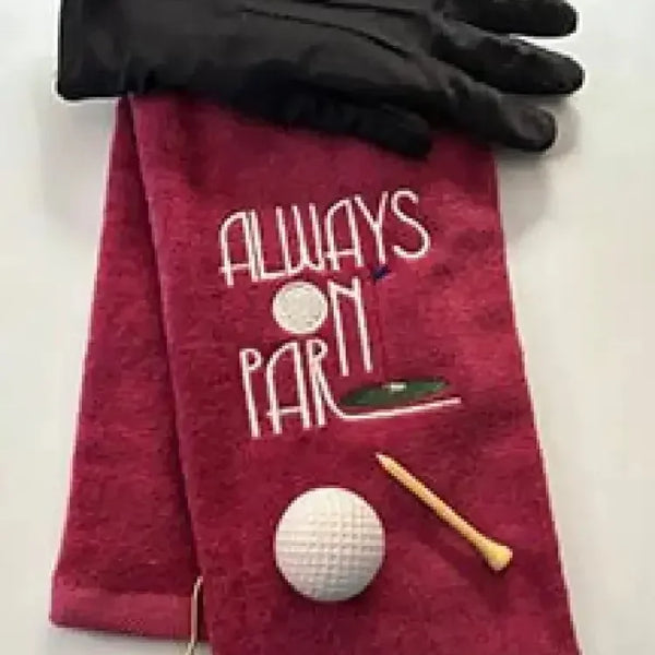 Red embroidered golf towel with 'Always On Par' text, black glove, golf ball, and tee