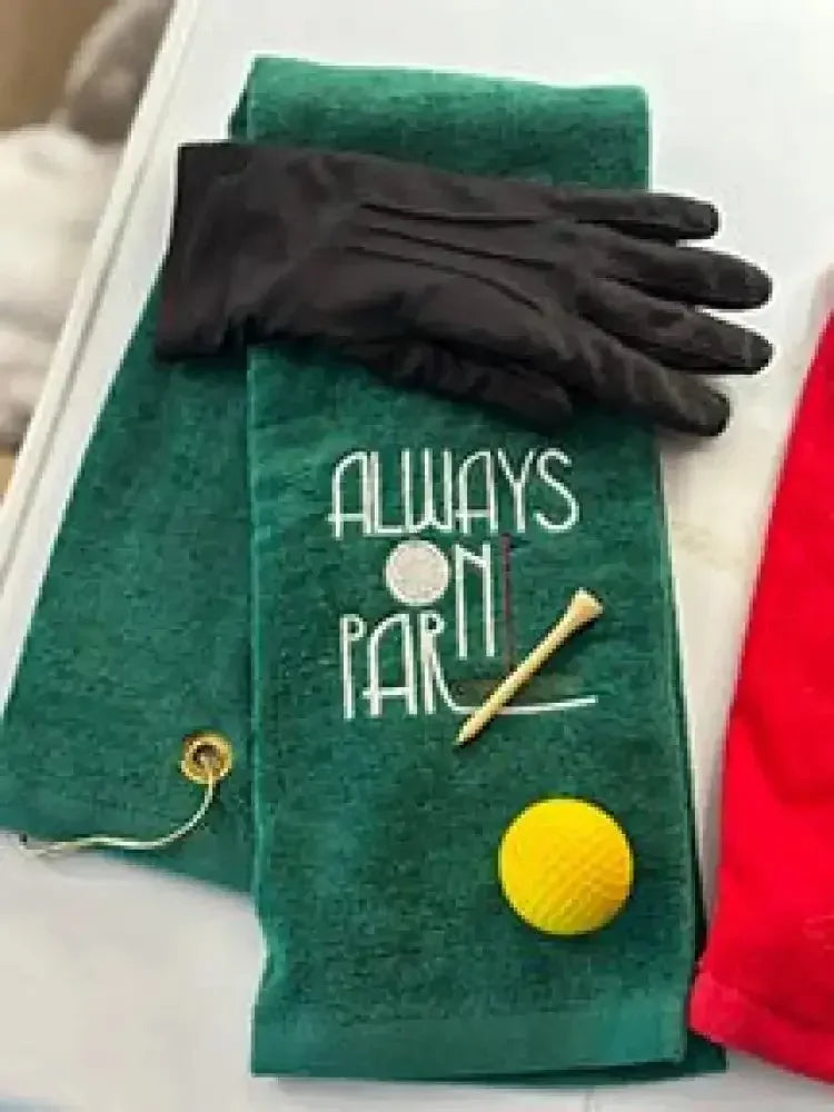 Green embroidered golf towel with 'Always On Par' text, black glove, tee, and yellow golf ball