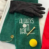Green embroidered golf towel with 'Always On Par' text, black glove, tee, and yellow golf ball