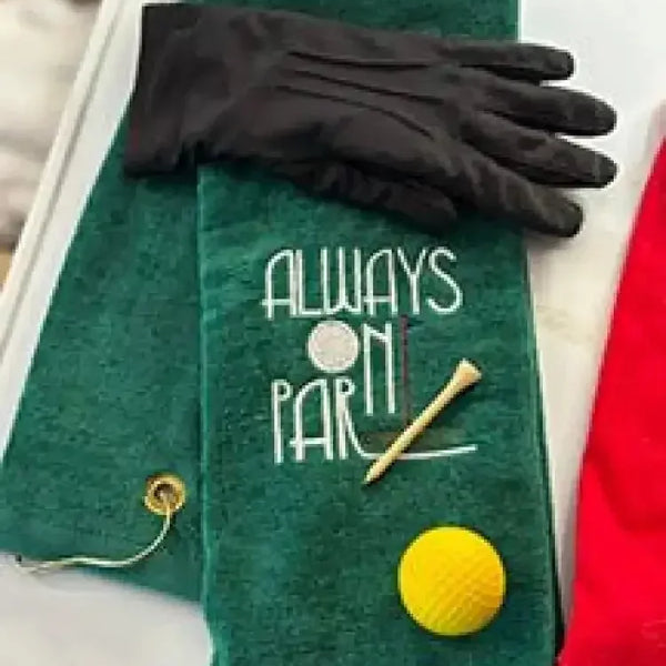 Green embroidered golf towel with 'Always On Par' text, black glove, tee, and yellow golf ball