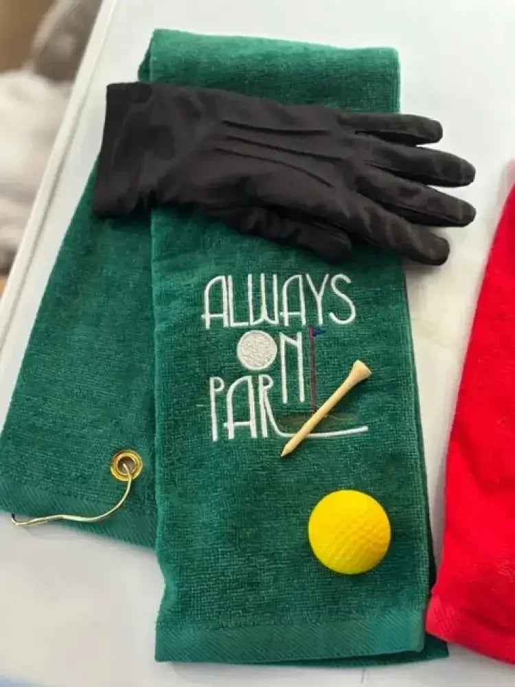 Green embroidered golf towel with 'Always On Par' text, golf glove, tee, and yellow ball