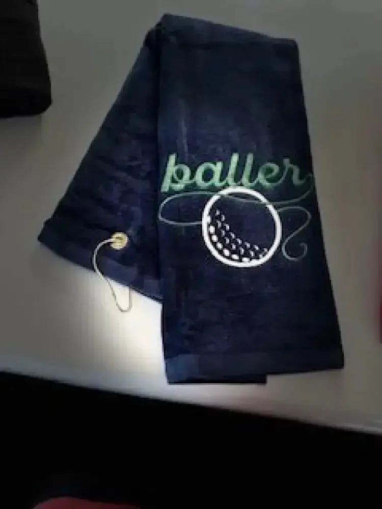 Navy blue embroidered golf towel with 'baller' text and golf ball design, metal grommet
