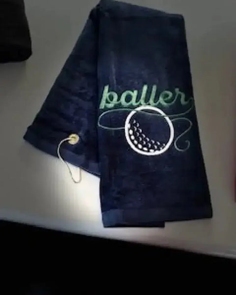 Navy blue embroidered golf towel with 'baller' text and golf ball design, metal grommet