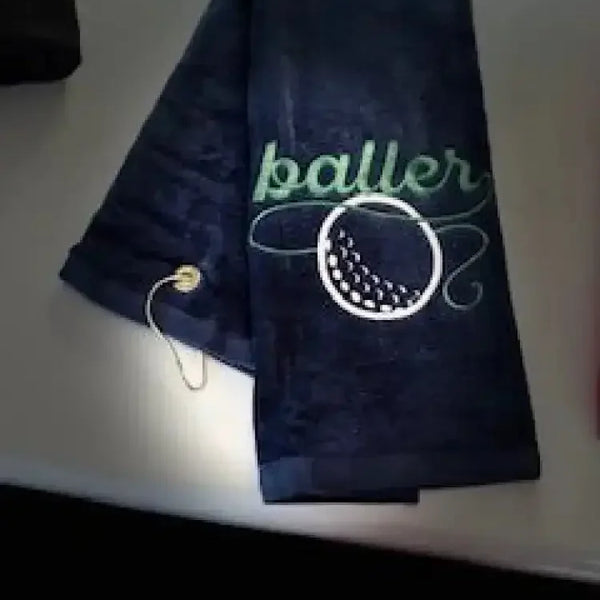 Navy blue embroidered golf towel with 'baller' text and golf ball design, metal grommet