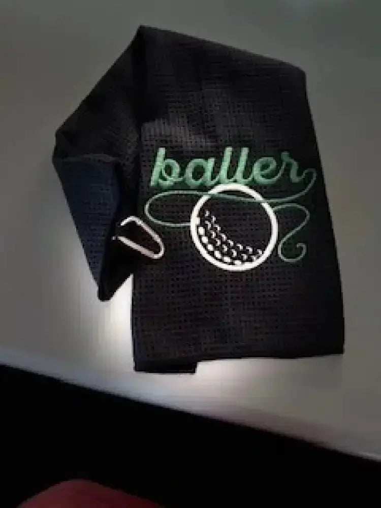 Black embroidered golf towel with 'baller' text and golf ball design, metal clip attached.