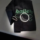 Black embroidered golf towel with 'baller' text and golf ball design, metal clip attached.