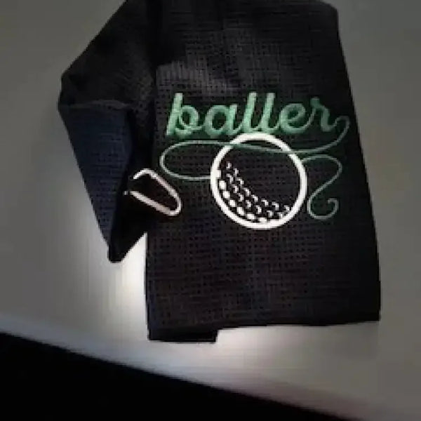 Black embroidered golf towel with 'baller' text and golf ball design, metal clip attached.