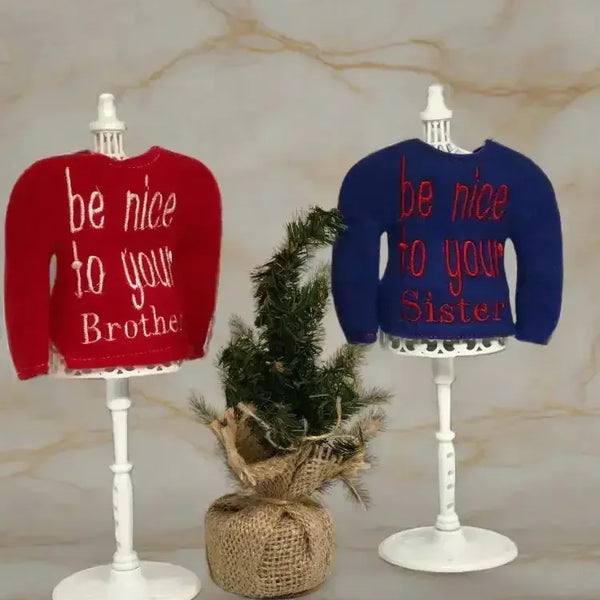 Red and blue embroidered elf sweaters on stands, with a small pine tree between them