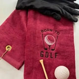 Red embroidered golf towel with 'Born to Golf' text, black glove, golf tee, and golf ball