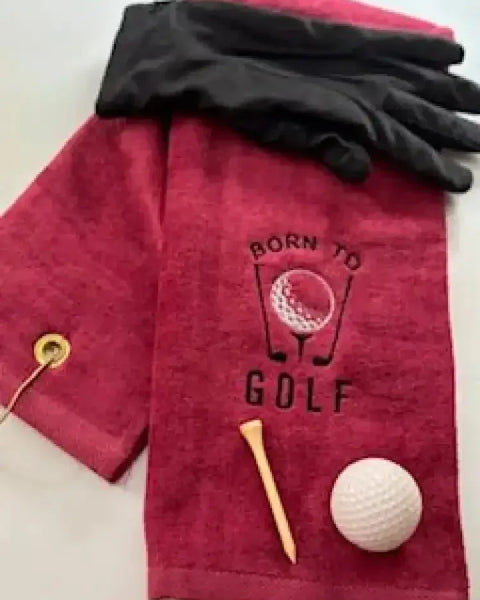 Red embroidered golf towel with 'Born to Golf' text, black glove, golf tee, and golf ball