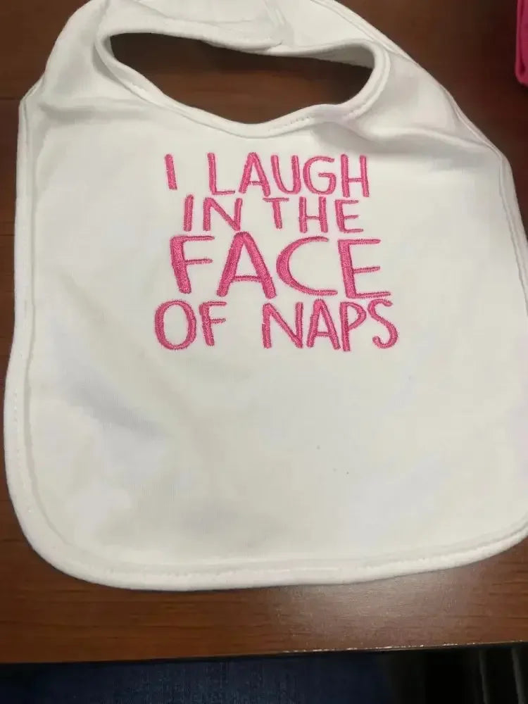 White baby bib with pink embroidered text saying 'I laugh in the face of naps'