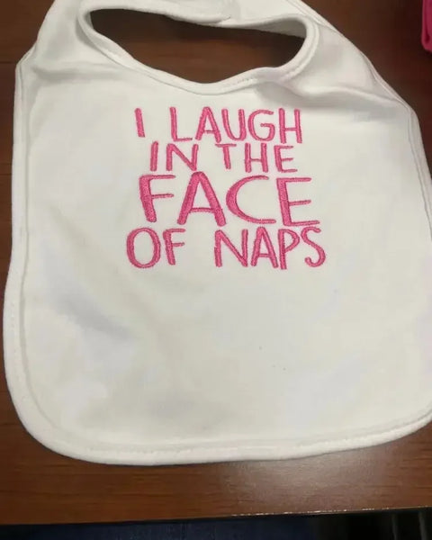 White baby bib with pink embroidered text saying 'I laugh in the face of naps'