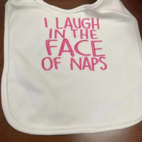 White baby bib with pink embroidered text saying 'I laugh in the face of naps'