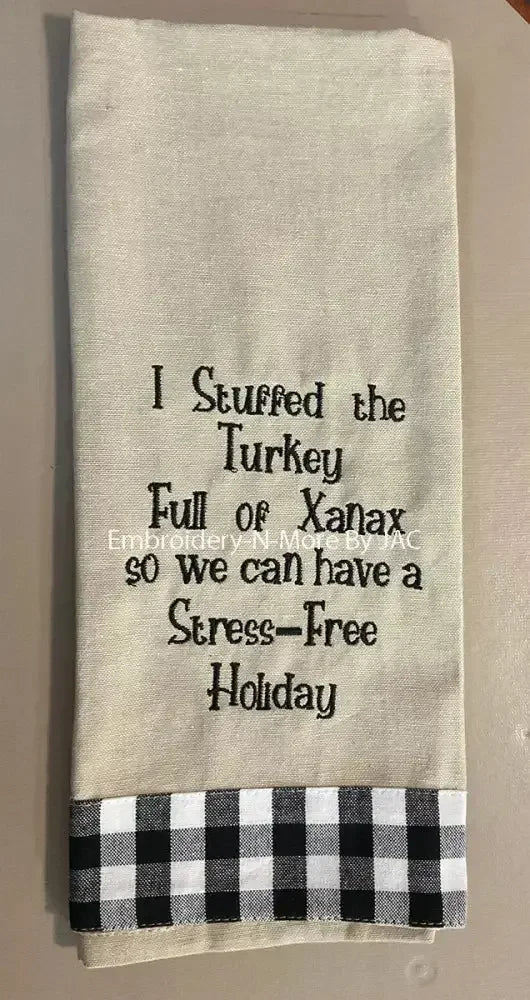 Beige embroidered hand towel with humorous holiday saying and black plaid trim.