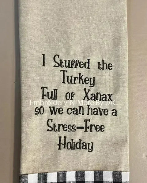 Beige embroidered hand towel with humorous holiday saying and black plaid trim.