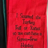 Red embroidered kitchen towel with humorous holiday text, waffle texture, black stitching.