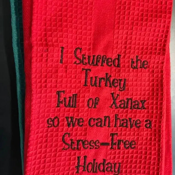 Red embroidered kitchen towel with humorous holiday text, waffle texture, black stitching.