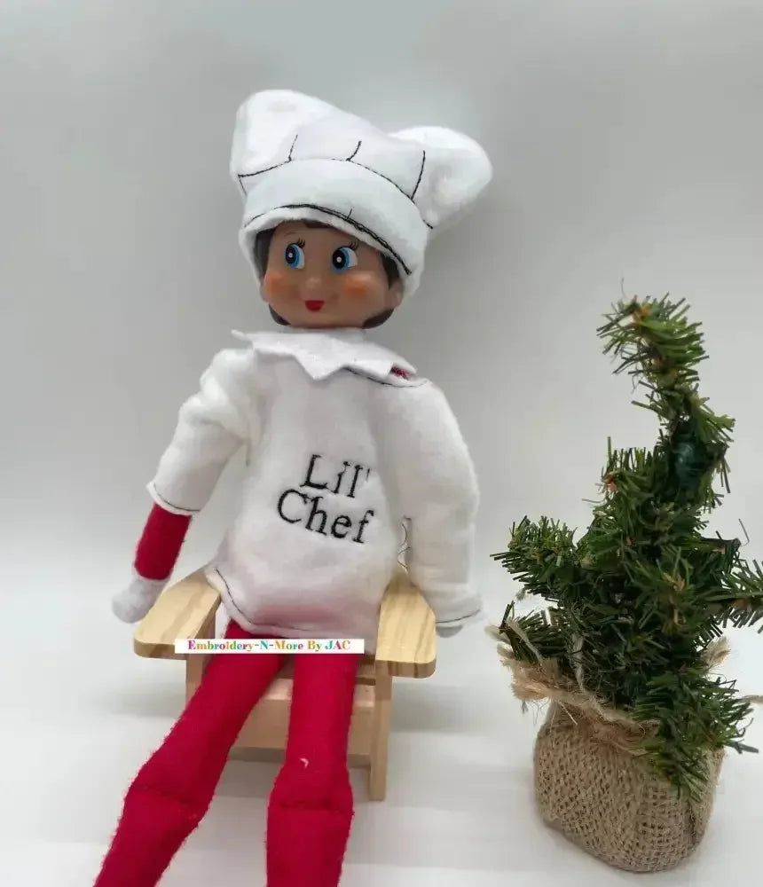 Elf doll wearing white chef costume and hat, sitting by a small decorative Christmas tree