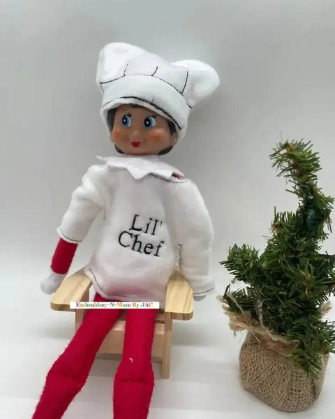 Elf doll wearing white chef costume and hat, sitting by a small decorative Christmas tree