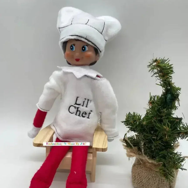 Elf doll wearing white chef costume and hat, sitting by a small decorative Christmas tree