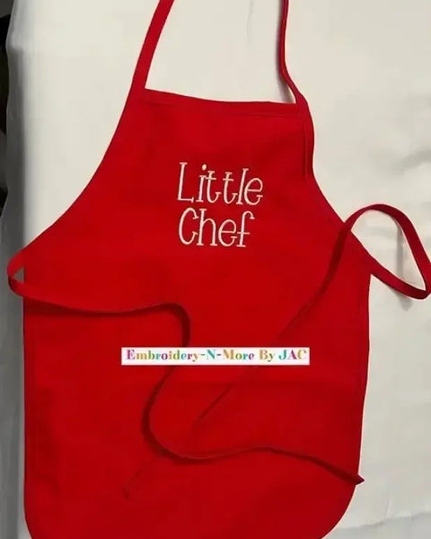 Red children's apron with 'Little Chef' embroidered on the front, displayed on a white surface.