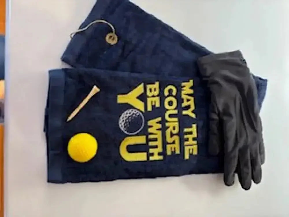 Navy embroidered golf towel with 'May the Course Be With You,' yellow golf ball, tee, and black glove.