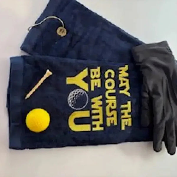 Navy embroidered golf towel with 'May the Course Be With You,' yellow golf ball, tee, and black glove.