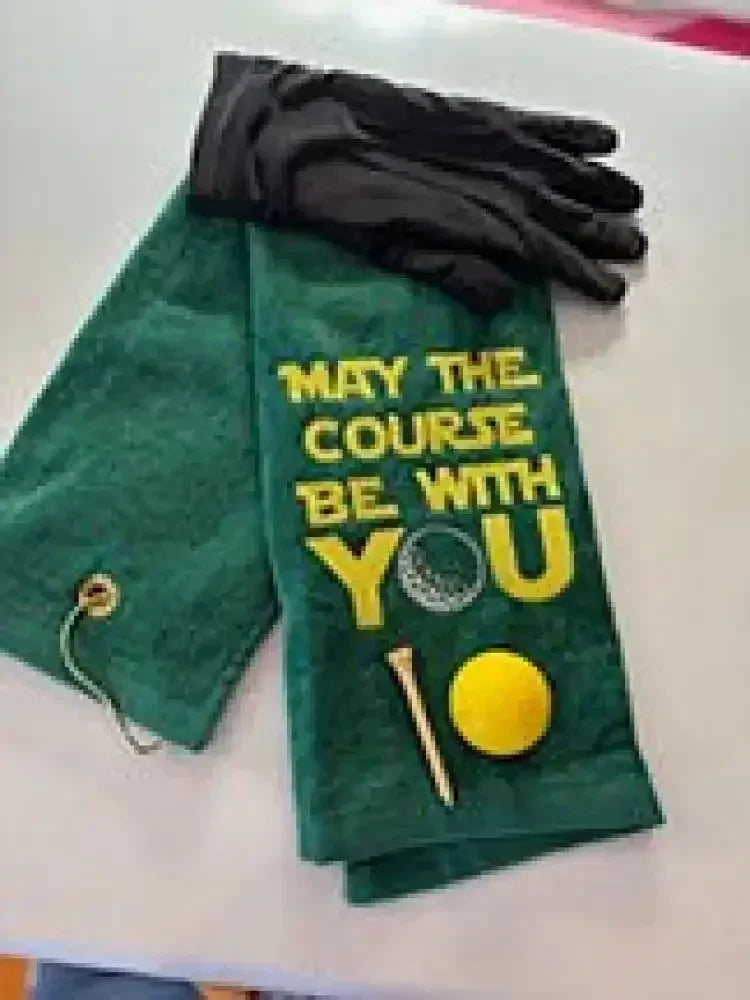 Green embroidered golf towel with 'May the Course Be With You' text, golf glove, ball, and tee.