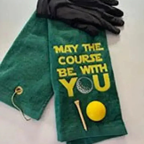 Green embroidered golf towel with 'May the Course Be With You' text, golf glove, ball, and tee.