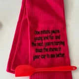 Red flour sack kitchen towel with humorous embroidered quote and red spatula on white surface