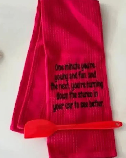 Red flour sack kitchen towel with humorous embroidered quote and red spatula on white surface
