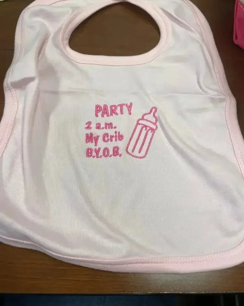 Pink embroidered baby bib with bottle and party-themed text on wooden table