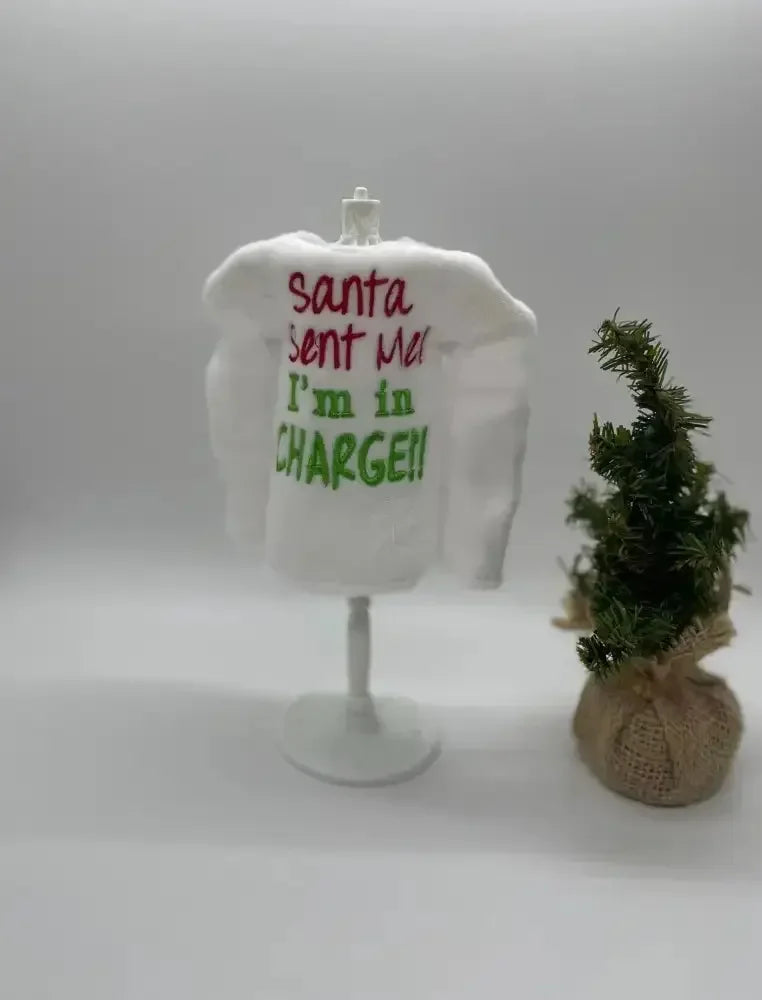 Mini white elf sweater with 'Santa sent me I'm in charge' embroidery beside small Christmas tree.