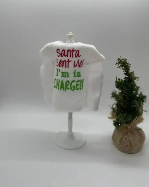 Mini white elf sweater with 'Santa sent me I'm in charge' embroidery beside small Christmas tree.