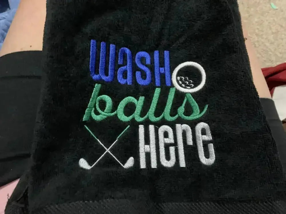 Black embroidered golf towel with 'wash balls here' text, golf ball, and crossed clubs design