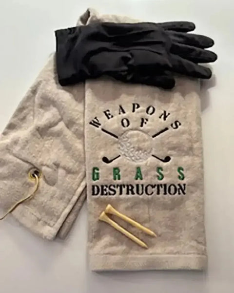 Embroidered golf towel reading 'Weapons of Grass Destruction' with golf gloves and tees