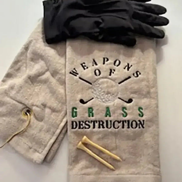 Embroidered golf towel reading 'Weapons of Grass Destruction' with golf gloves and tees