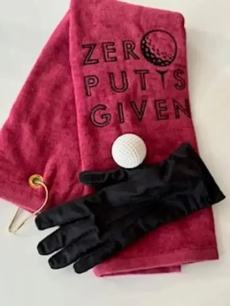 Red embroidered golf towel with 'Zero Putts Given,' golf ball, and black glove on white background.